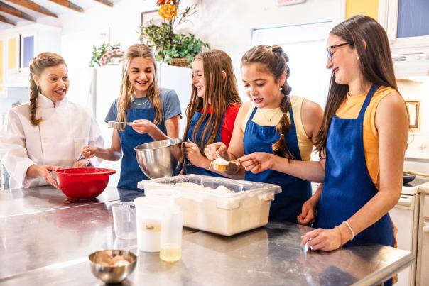 Four teen girls in blue aprons and a cooking instructor in a white smock stand at a kitchen prep table measuring cups of flour from a large, rectangular tub.