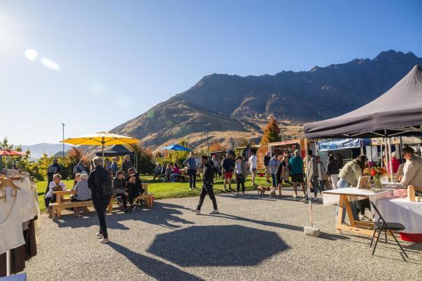 People browsing stalls and relaxing under colourful umbrellas at the Remarkables Market, with mountains in the background on a sunny day.