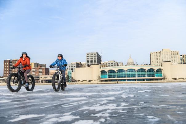 Two people, one in an orange jacket, one in a blue jacket, riding bicycles with fat tires on a frozen Lake Monona with Monona Terrace and the Capitol in the background