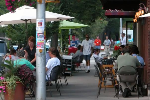 A bustling scene on Rochester's Park Avenue. Featuring outdoor dining at Jine's Restaurant, with pedestrians and vibrant flower planters lining the sidewalk.