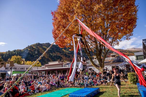 Crowd gathered around an aerial performance sounded by red and gold trees at the Arrowtown Autumn Festival