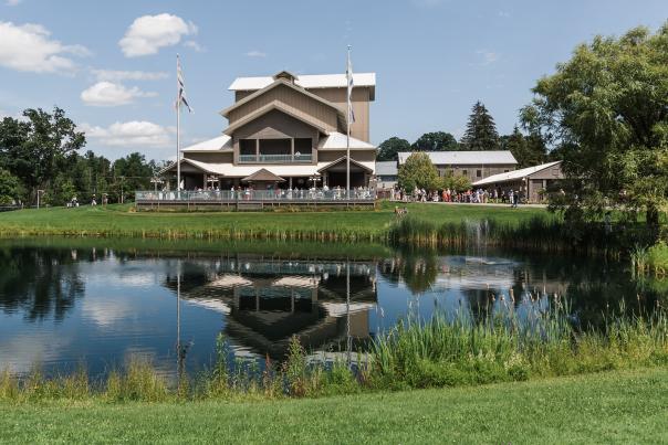 The outside of a theater building with its reflection showing in the pond in front of it. The pond and theater are surrounded by green grass and trees and there are people on the deck in front of it.