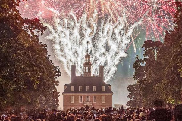 Fireworks illuminate the night sky above a historic building, surrounded by a crowd of spectators. Lush trees frame the scene, enhancing the festive atmosphere.