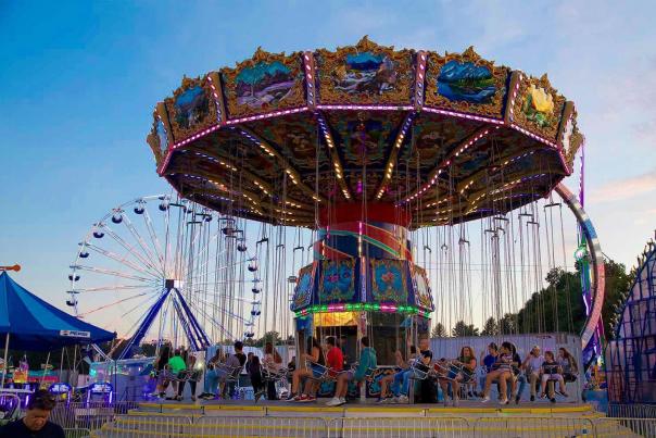 Festivalgoers sit aboard a brightly lit swing ride with a Ferris wheel in the background at Slidell Food & Fun Fest.