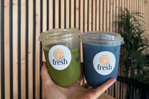 Hand holding up two Fresh juice drinks in front of a logoed wall.