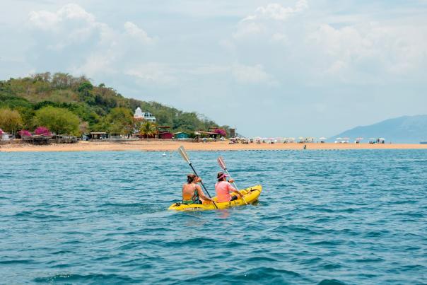 Two individuals paddle a yellow kayak in the blue waters near a tan sandy beach and a green, hilly island under a partly cloudy sky.