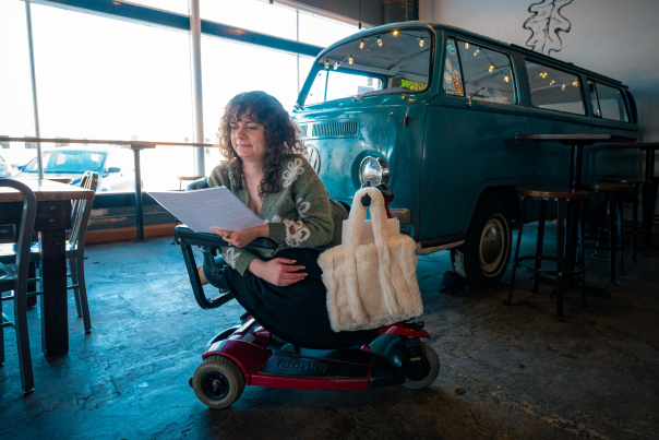 A woman on a mobility scooter reads a document in a cozy cafe, near a vintage blue van. Warm lighting, soft expression, and casual ambiance.