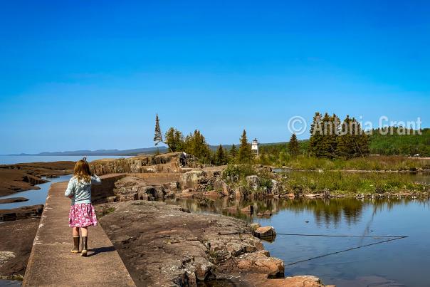 Young girl in pink polka dot skirt walking on the narrow concreate breakwall on the Grand Marais harbor with the lighthouse visible in the background.