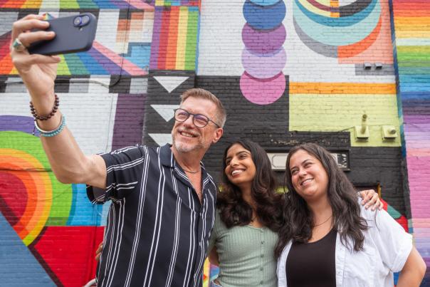 Three people pose in front of a colorful mural while they take a selfie.