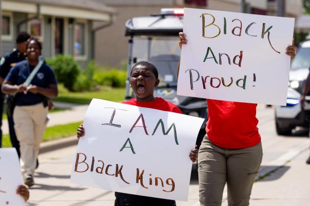 A young Black boy yells while marching in a Juneteenth parade. He is holding a sign that says "I Am a Black King." Behind him, a person is holding a sign that says "Black and Proud."