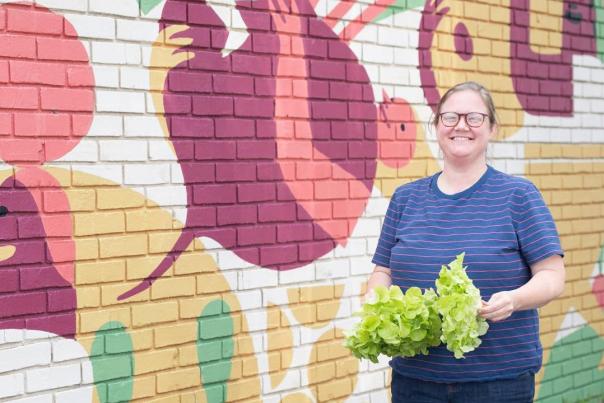 Mimi Maumus of home.made stands in front of a colorful mural, holding vegetables.