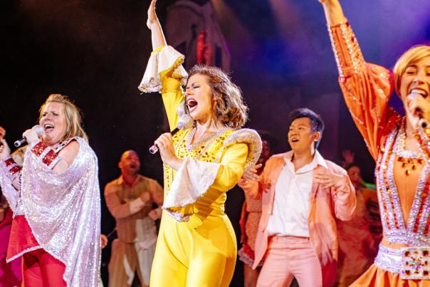 Three women dressed up in 70s outfits sing on stage with their hands raised in unison. Behind the three women is a man dancing backup. The group is singing a song.