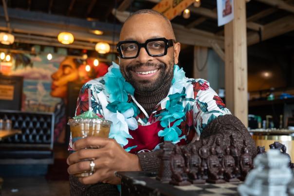 Man wearing thick black glasses sits at a chess table and smiles at at the camera, while holding an iced coffee in a plastic cup.