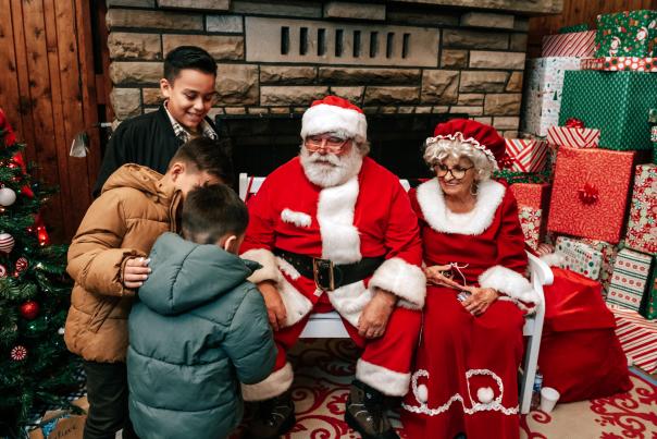 Santa and Mrs. Claus sitting on a bench inside a holiday village building talking with three young children. The building is decorated with Christmas decor,  a tree, and wrapped presents