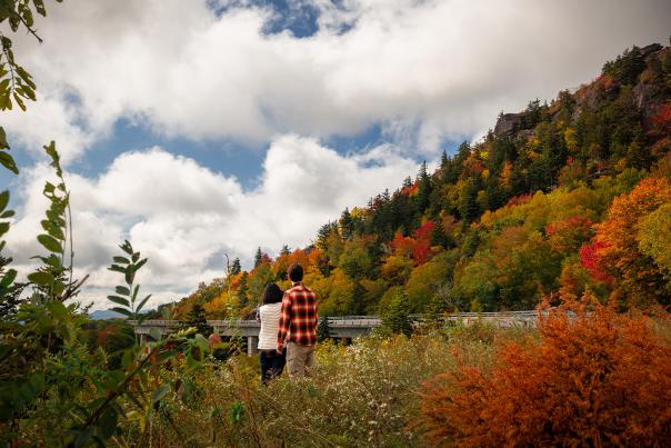 A woman in a white sweater and a man in red flannel view the beautiful fall foliage from the side of the Blue Ridge Parkway.