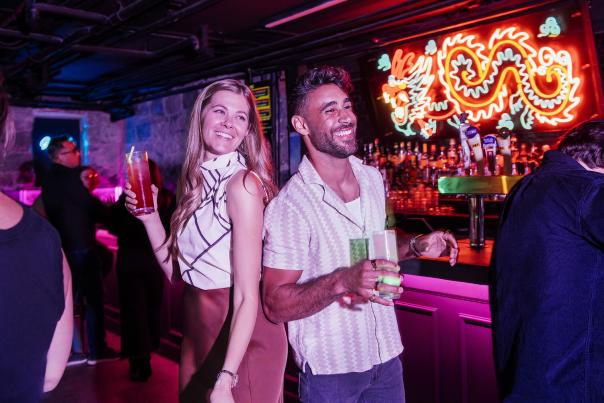 Couple with cocktails in underground bar with neon dragon sign behind