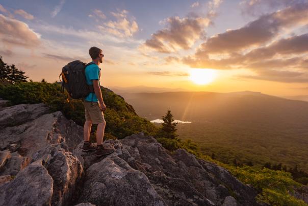 A man wearing a large backpack looks out over a sunset from Grandfather Mountain's peak.
