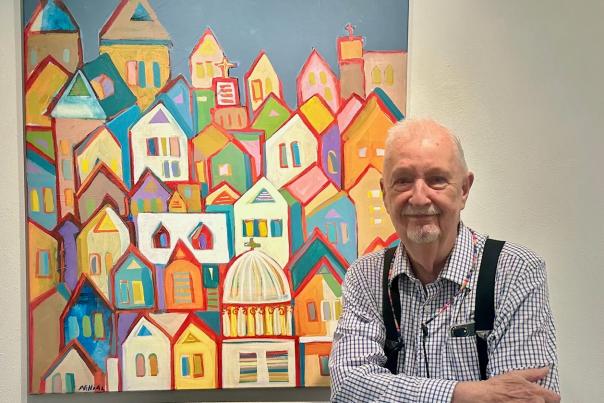 A white-haired man in a white-checkered shirt (Jim N. Hill) stands in front of one of his paintings displayed in TWAC's exhibit space. The painting depicts a colorful skyline of overlapping houses and churches of various faiths.