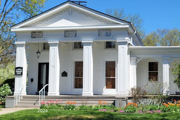 white building with a greek revival architectural style sits on a green grassy site.