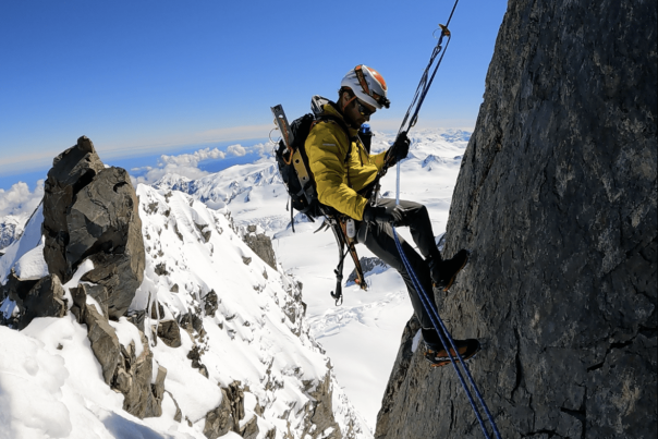 Mountaineer Hamish Fleming rappelling down a rock face with snow-covered mountains in the background