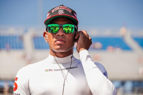 Indy NXT driver Yuven Sundaramoorthy stands at a racetrack wearing a Wisconsin cap, mirrored sunglasses and a white fire-resistant racing shirt while adjusting an earpiece. His sunglasses reflect activity on the track, with blurred grandstands and blue stadium seating visible in the background.