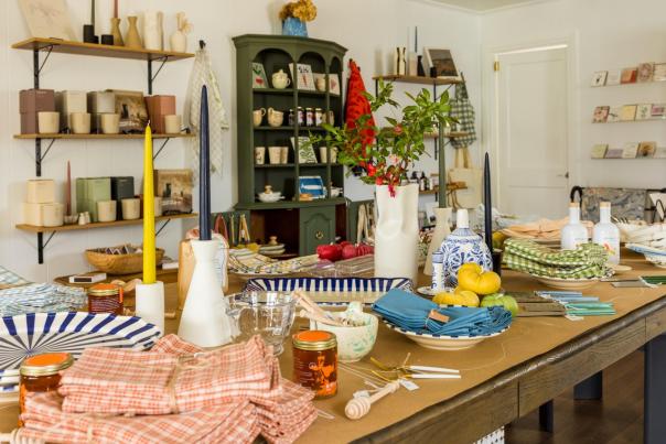 A cozy shop interior featuring a wooden table set with colorful tableware, candles, and fresh produce. Shelves display various home goods and decor.