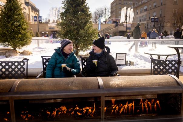 Two women in winter clothes chat while drinking warm beverages in front of a fire outside at The Edgewater Hotel ice rink