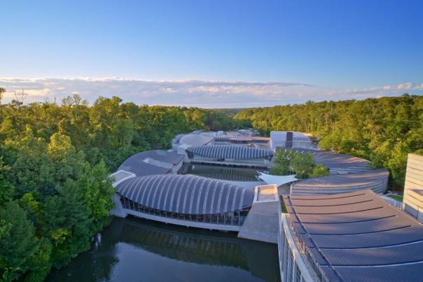 Aerial view of modern architectural structures nestled among lush greenery, reflecting on a serene waterway under a clear blue sky.