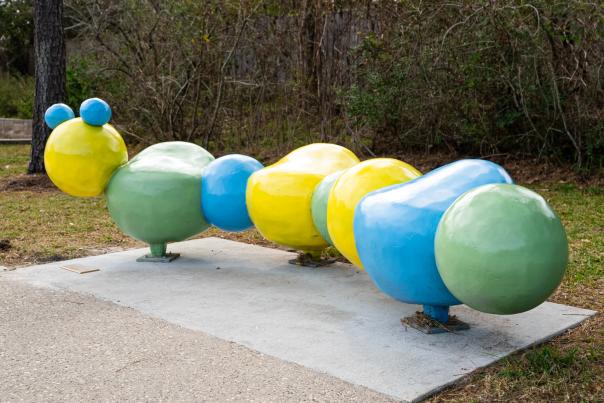 An art bench looks like a caterpillar out of a children's book. Its body is composed of eight rounded body segments in various sizes, each alternating in colors of bright yellow, green, and blue. Two blue spheres sit atop its head, representing the creature's eyes.