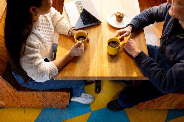 A photo from above of a woman's and a man, both holding coffee, at a table in a coffee shop next to a laptop