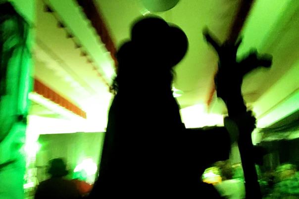 Silhouette of man with hat on walking around in room during a theatre production. The room is filled with green lighting, creating a haunting or halloween-themed tone.