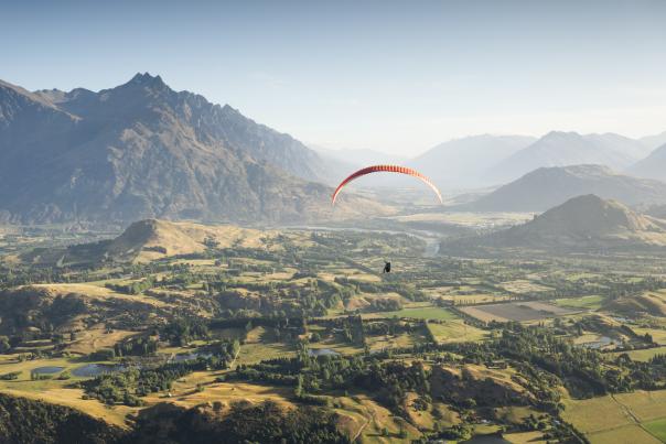 Person paragliding over a green valley with the Remarkables mountain range in the background