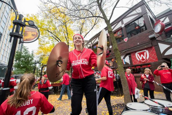 A white woman from the UW Marching Band holds cymbals on a Fall day outside of State Street Brats