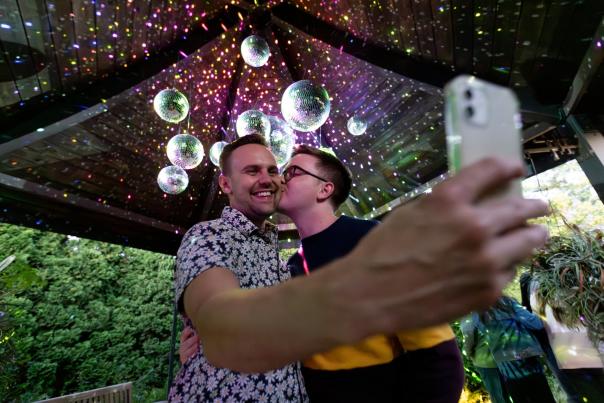 Two men stand under disco balls and take a selfie while one man kisses the other man on the cheek