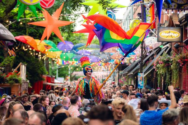 Person dressed in Pride colours waving flag above crowd on Canal Street, Manchester