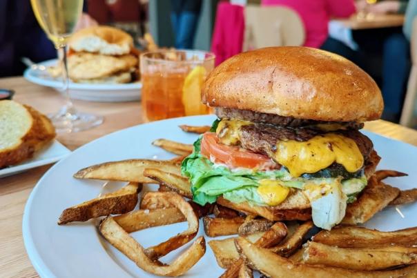 A large burger with cheese, tomato, lettuce and sauce served on a white plate with fries. An old fashioned and glass of champagne are also on the table.