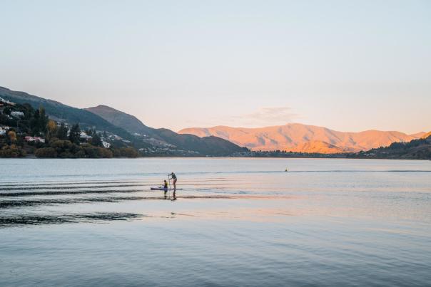 Paddle boarder in middle of still lake at dusk with mountains in the background
