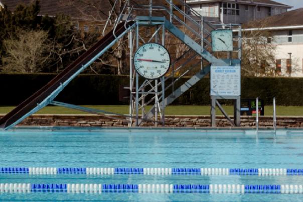 An image of Cheltenham Lido with the water in the foreground and in the background a set of steps with a big timer on it and a slide coming off the side.