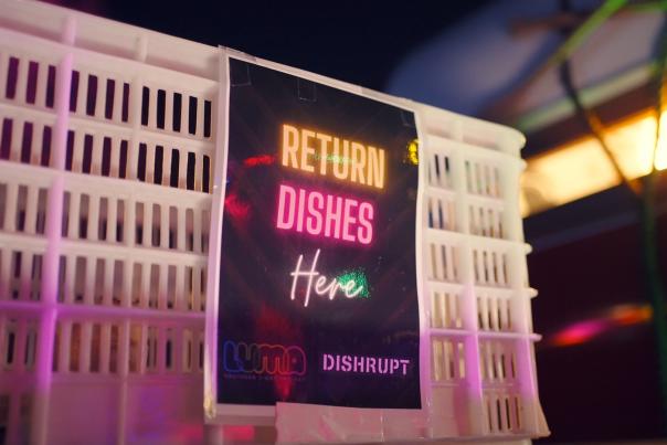 Sign illuminated by different coloured lights that read Return DIshes Here, LUMA x DISHRUPT