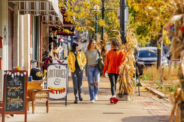 Three women shopping in Depot Town, walking along storefronts on a sidewalk during the fall season. Yellow and orange leaves in the background.