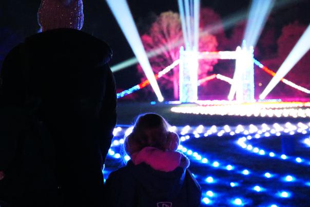 A mother and small child stand with their backs to the camera looking at a small, lit up London Bridge. There are roaming spotlights & glowing fairylights on the floor in all different colours. The trees off in the distance are also lit up in blue and pink.