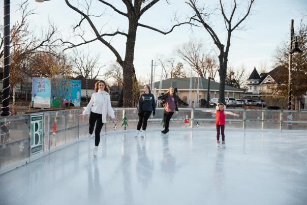 Four people skate on an outdoor ice rink surrounded by trees and buildings. Colorful decorations hang around the rink, enhancing the festive atmosphere.