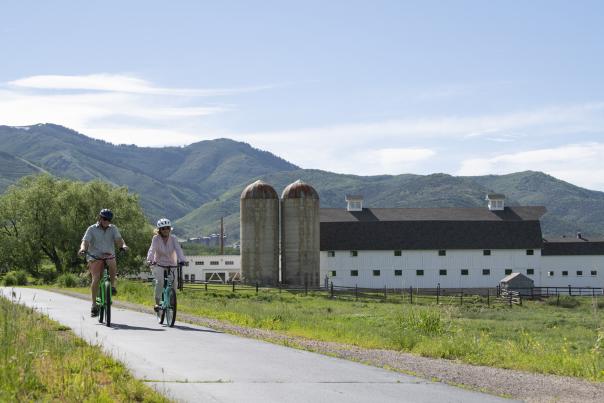 Two people biking a paved trail with the white McPolin Farm in the background.