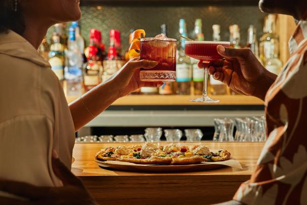 A couple shares drinks over a pizza at the True Food Kitchen bar. The handcrafted pizza features a thin crust, spinach, peppers, and large balls of melted cheese.