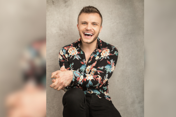 a man sat against a neutral background smiles at the camera with his hands together
