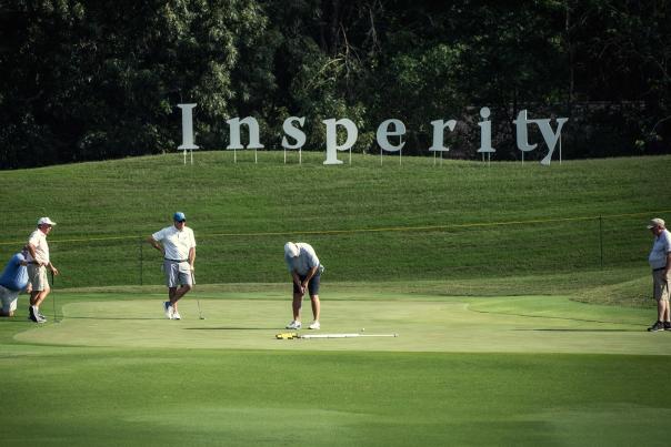 A group of four men, 50s-60s, watch a fifth man putt a golf ball into the hole. On the grassy hill behind them are large letters spelling "Insperity."