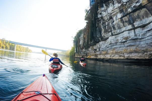 View from kayaking of person kayaking ahead on TN river, cliff to the side and bridge in front