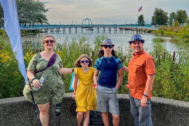 Four family members posing by a lakeside with a bridge in the background under a clear sky.