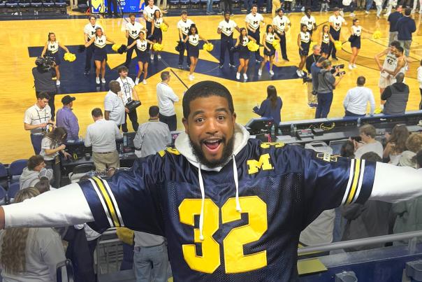 Man expresses excitement with his arms outstretched, wearing a jersey with the number 32 on the front. Behind him is a basketball court with University of Michigan cheerleaders performing on the court.