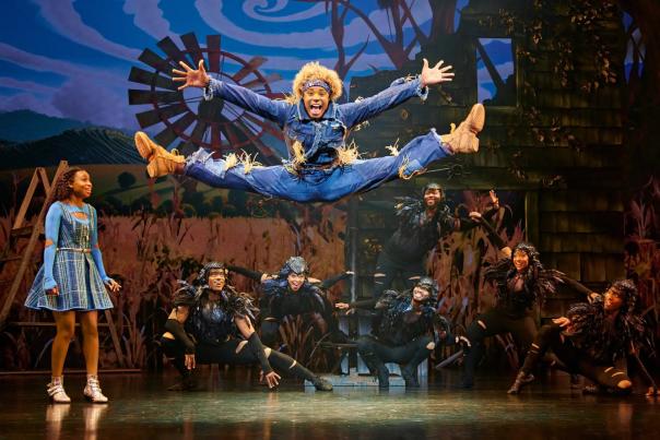 A performer dressed as the Scarecrow leaps high into the air with legs spread wide in an energetic dance move, while another actor dressed as Dorothy watches from the side. The stage background features a windmill, cornfield, and swirling sky, creating a lively and colorful scene from The Wiz.